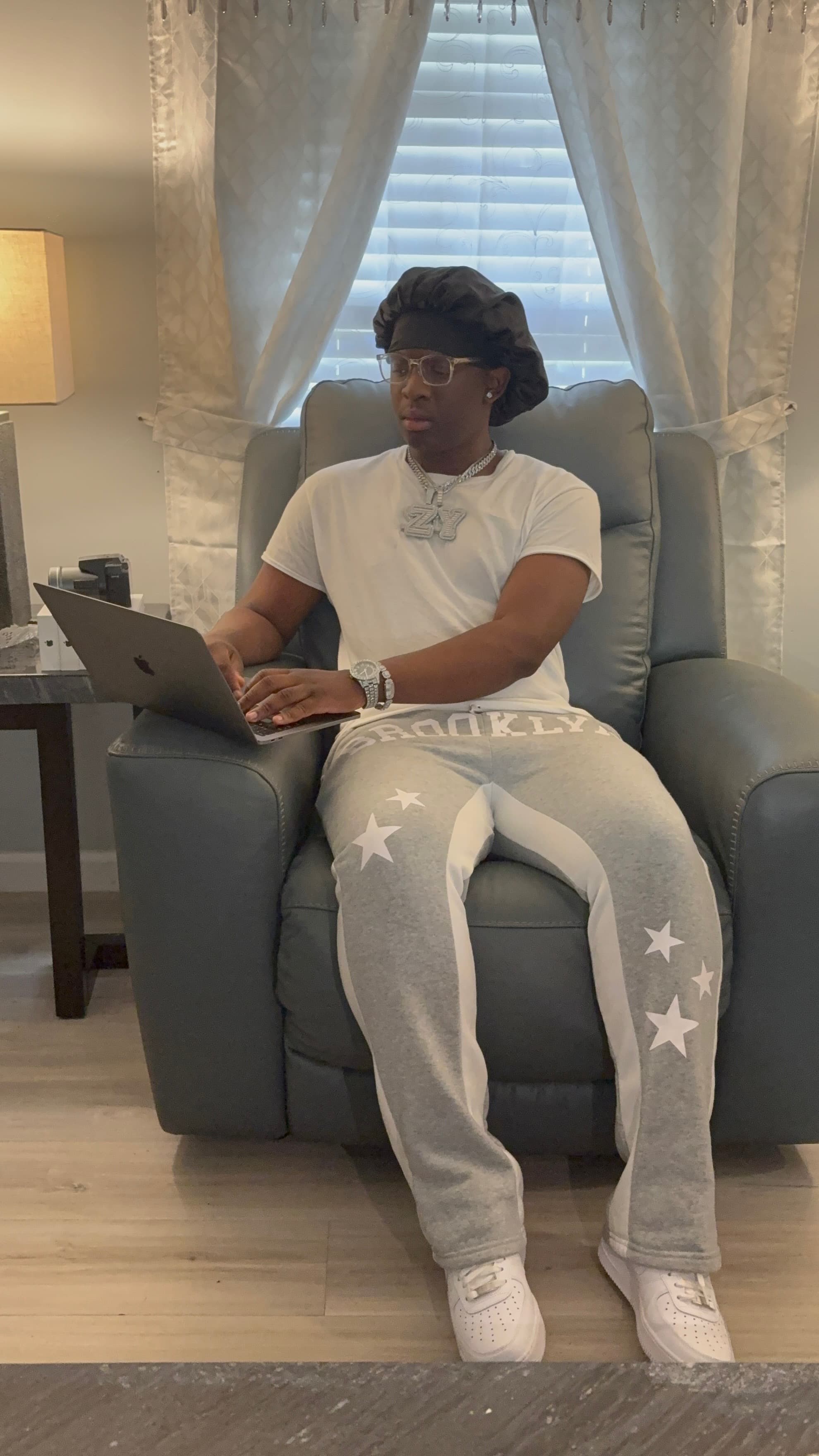 Young man wearing a bonnet and jewelry sits in a grey armchair using a laptop.